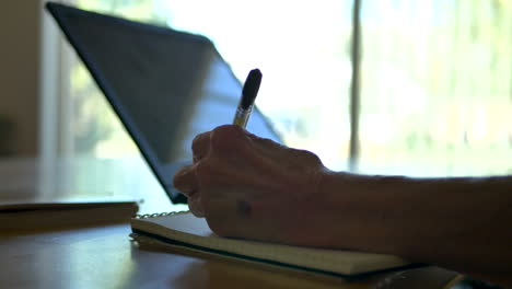 an elderly woman with wrinkled hands writes with a pen in a notebook as she look on her laptop backlit by a bright window