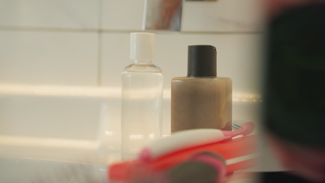 Gæstens hånd rækker ud efter en flaske med toiletartikler på hotelvasken ved siden af en rød tandbørste; fingre løfter låget og placerer flasken under vandhanen; varm, flisebelagt baggrund med diskret damp, nærbillede af gæstfrihedens detaljer.