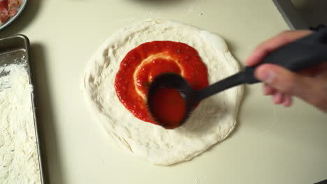 vista superior del chef mientras esparce salsa de tomate sobre masa de pizza con un cucharón en el restaurante de pizza italiana tradicional
