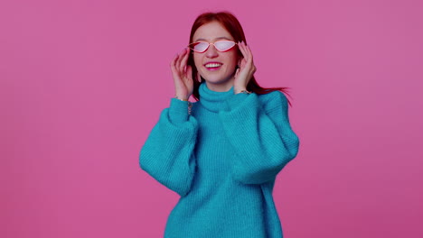 seductive cheerful redhead girl in blue sweater wearing sunglasses, charming smile on pink wall