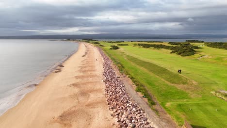 drone footage glides above a sandy shoreline and adjacent golf course in rosemarkie, highland, scotland, under overcast skies with soft, natural daylight