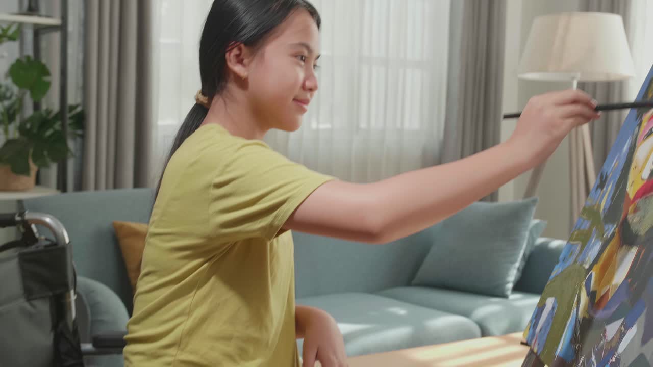 Side View Of An Asian Artist Girl In Wheelchair Holding Paintbrush And Painting A Girl On The Canvas