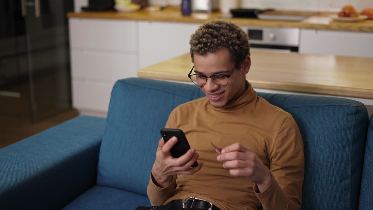 joven feliz comprando en línea usa un teléfono y una tarjeta de crédito en el sofá de casa