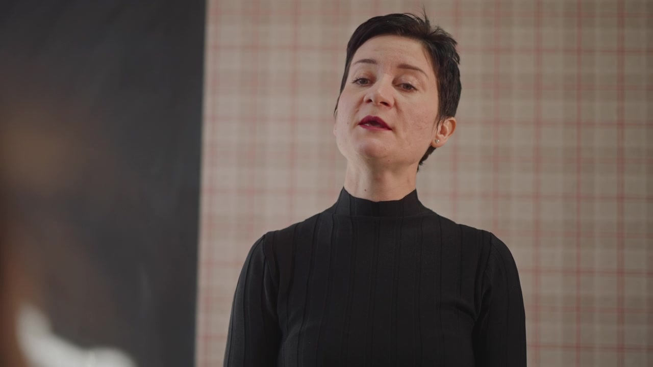 Choir conductor leading rehearsal in classroom, shorthaired instructor projects voice and engages offcamera students, confident posture and encouraging expression, chalkboard and plaid wallpaper