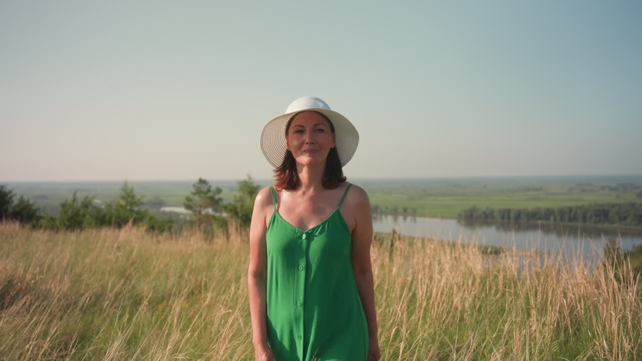 Donna in abito verde e cappello da sole bianco cammina con calma tra l'erba alta e dorata su un pendio soleggiato con un sorriso dolce, mentre una lussureggiante valle verde e un fiume si estendono dietro sotto un cielo limpido.
