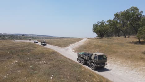 israel army squad soldiers on heavy vehicles driving through green field at training ground country road, aerial shot