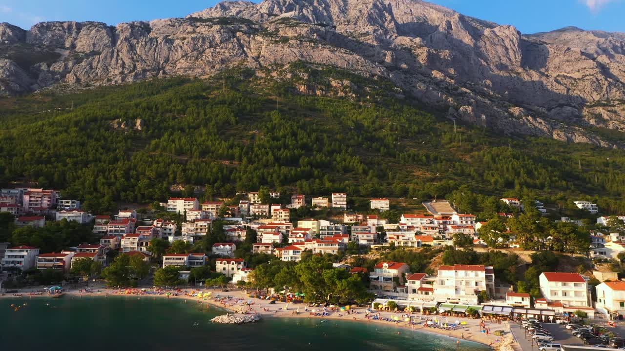 Widok z lotu ptaka oddalający się od plaży, odkrywający ogromną górę i morze podczas zachodu słońca w Baškiej Vodzie, Chorwacja, w jakości 4K.