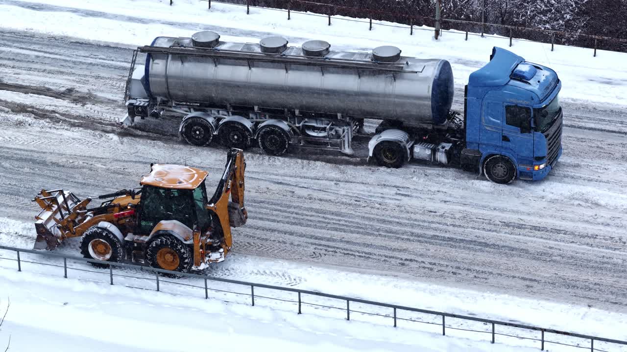 Drone aerial of a blue fuel tanker stuck on an icy winter roadway while a snowplow backhoe works nearby, highlighting hazardous traffic conditions and road maintenance