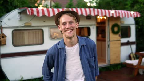 Portrait-Of-Happy-Handsome-Bearded-Man-Wearing-Denim-Jacket-And-White-T-Shirt-And-Hat-Smiling-And-Looking-At-Camera-While-Standing-Near-House-On-Wheels-Outdoors