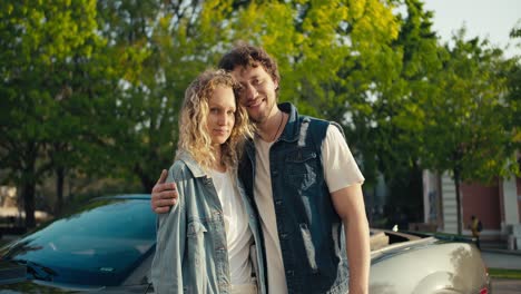 Portrait-of-a-young-brunette-guy-and-a-blonde-girl-who-are-standing-near-their-gray-car-in-denim-jackets-smiling-and-looking-at