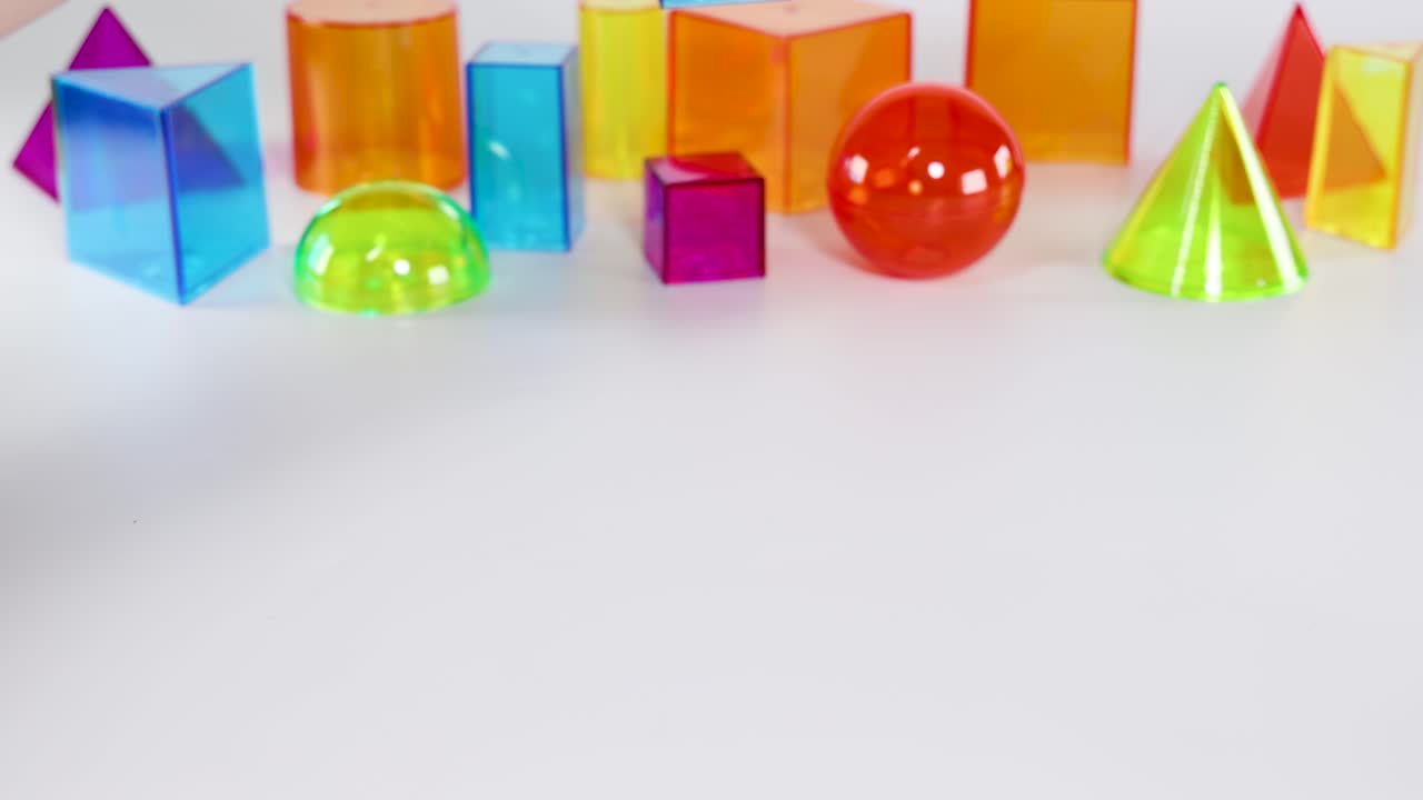 Hands interact with vibrant 3D geometric shapes on a white surface, illustrating basic mathematical concepts in a playful setting