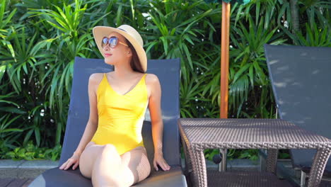 an attractive young woman in a yellow bathing suit, sunglasses, and straw hat sits poolside in a sun lounger