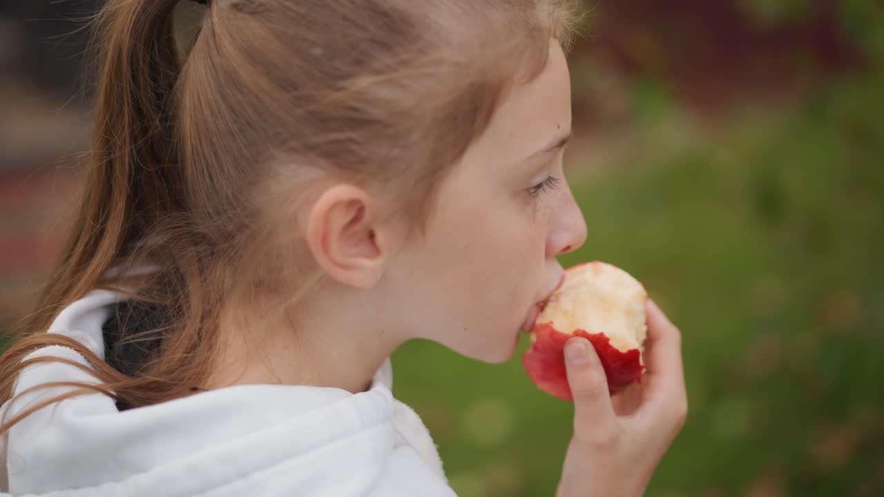 Young white girl eating apple outdoors in suburban backyard, white hoodie, ponytail, closeup profile, taking slow bites, autumn foliage background, candid family lifestyle moment, wholesome snack