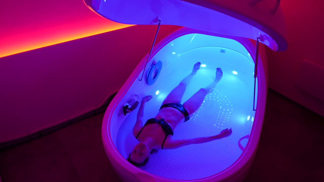 Woman float in relaxing hyper-saline floatation therapy pod in pink lit room