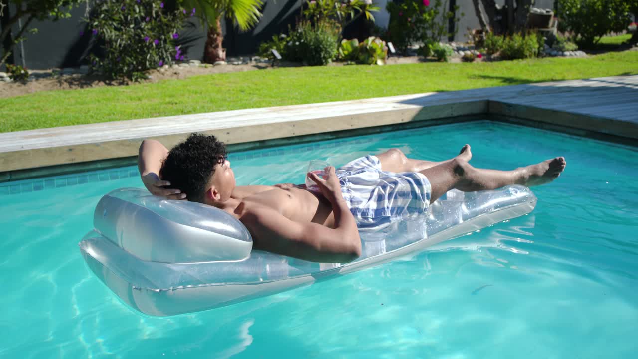 Man in swim-trunks lying on clear-float in pool with cell, camera pulling back showing yard