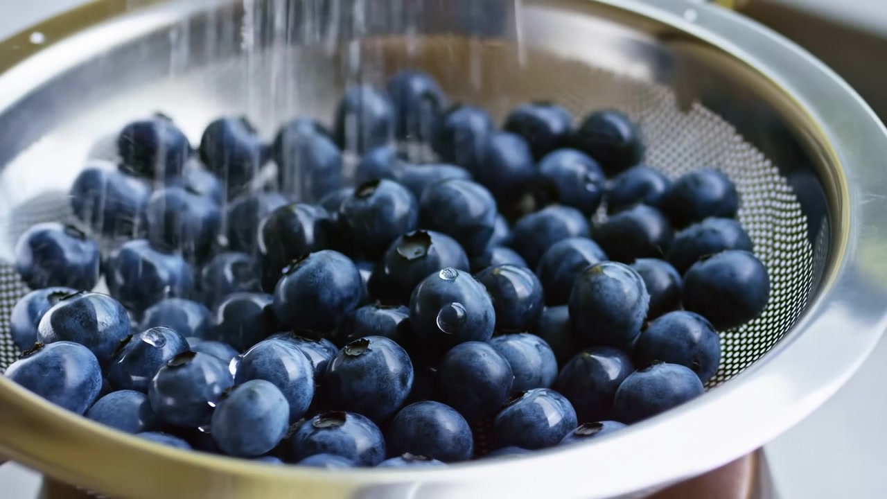 Close-up de mirtilos escuros sendo enxaguados sob água corrente em uma pia de cozinha, com luz natural brilhante e um ambiente limpo de preparação de alimentos.