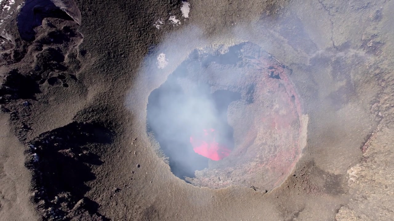 Aerial view of a volcanic caldera revealing bright red lava bubbling beneath a thin layer of smoke.
