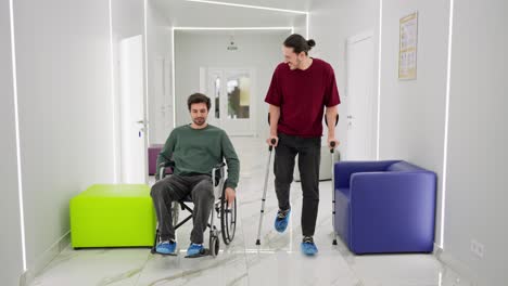 un hombre moreno feliz con paja en una chaqueta verde monta en una silla para discapacitados durante su rehabilitación y se comunica con un hombre con una camiseta roja que está caminando con muletas en una clínica moderna