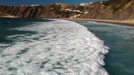 Luftdrohnenaufnahme-Von-Surfern,-Die-Im-Wasser-Liegen-Und-Neben-Einigen-Häusern-Auf-Einer-Klippe-In-Der-Algarve,-Portugal,-Durch-Wildwasser-Schwimmen