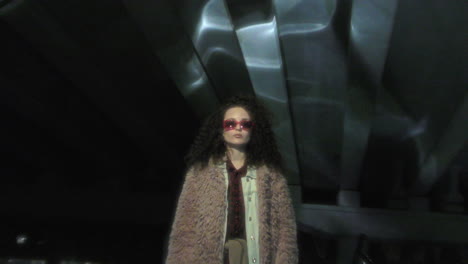 portrait of young woman in sunglasses and fur coat under dark concrete overpass