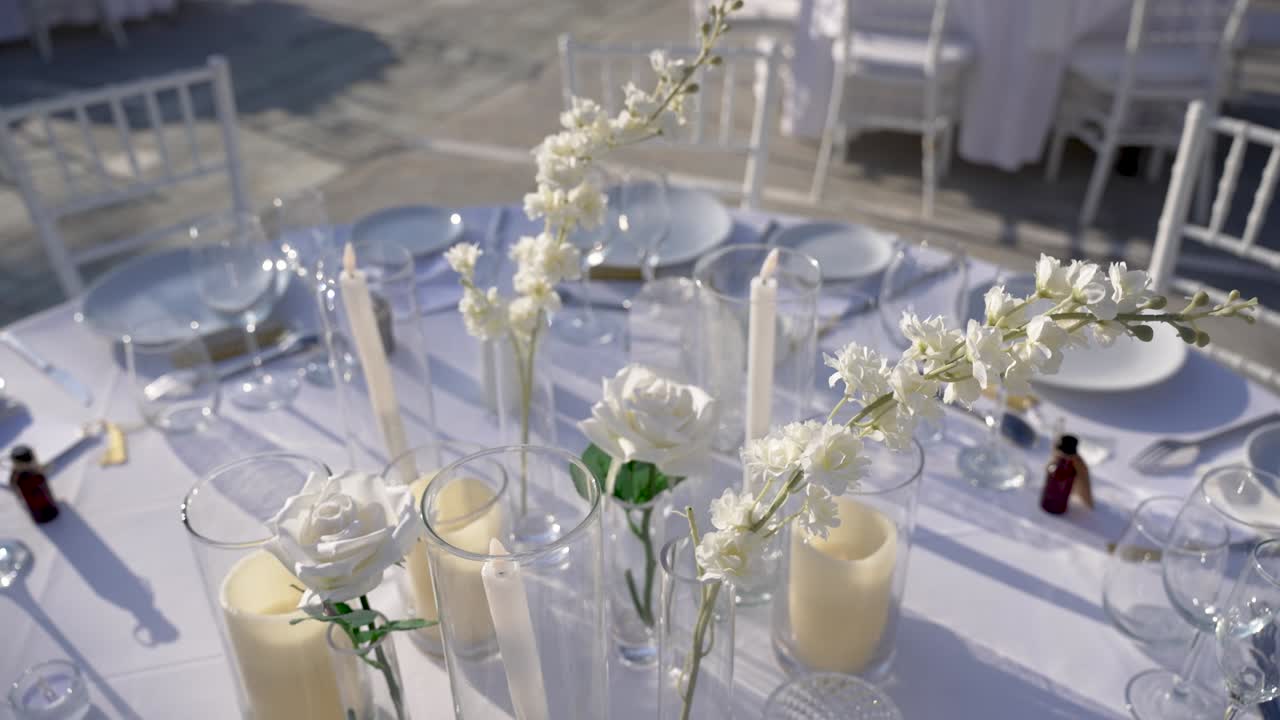 Un tavolo è imbandito con fiori bianchi e candele in alti bicchieri. Piatti e bicchieri sono disposti in preparazione di un evento all'aperto sotto la luce intensa del sole.