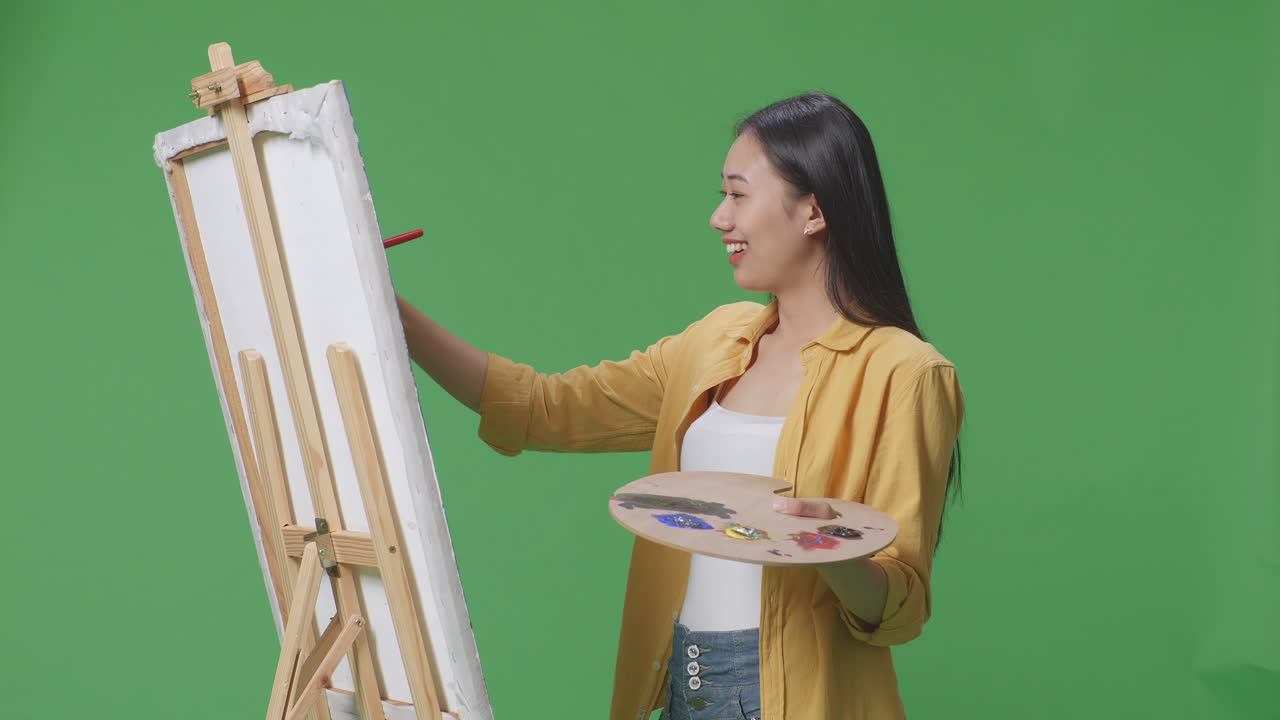 아시아 여성 예술가가가 팔을 고 그림을 보면서 캔버스에 기름색 페인트와 질로 그림을 그리고 있습니다.