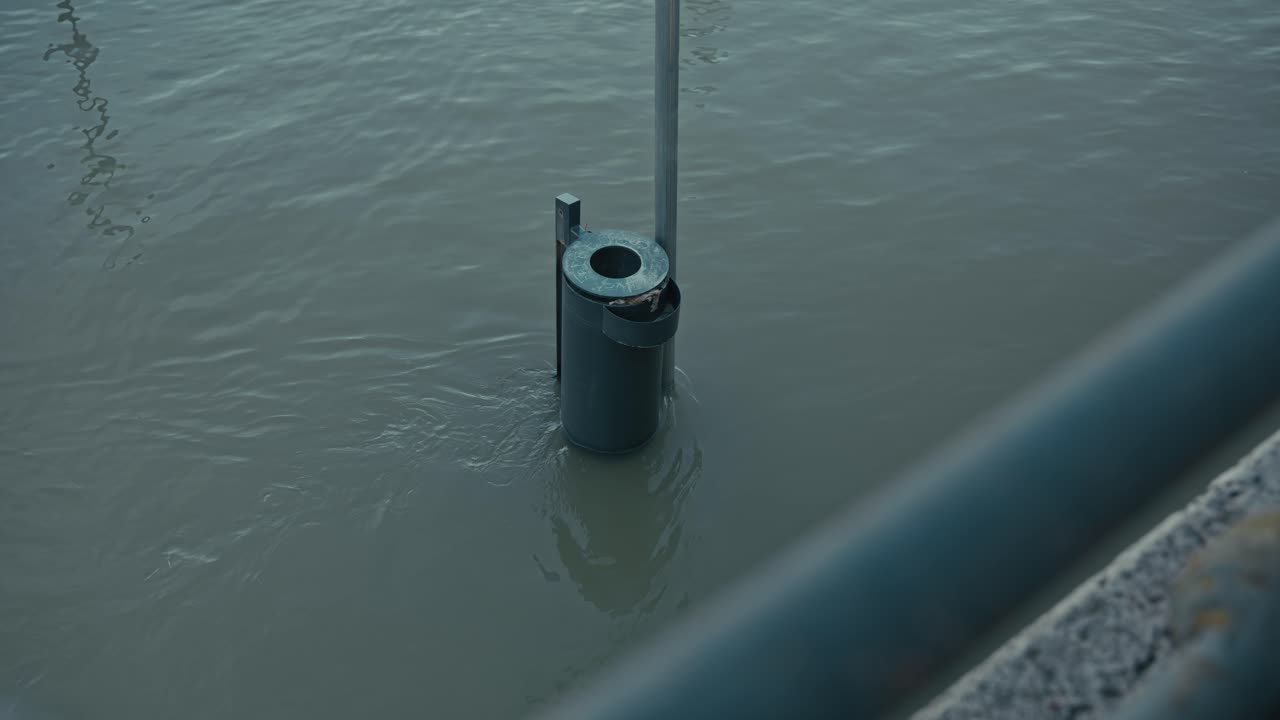 trash bin partially submerged in floodwaters near a pole, with calm water surrounding it, Budapest, Hungary