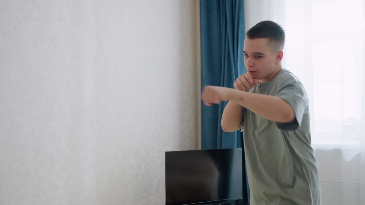 Teen kick boxer in loose shirt and cargo pants stands with fists raised practicing punches inside bright modern living room, near curtain window and displaying focus