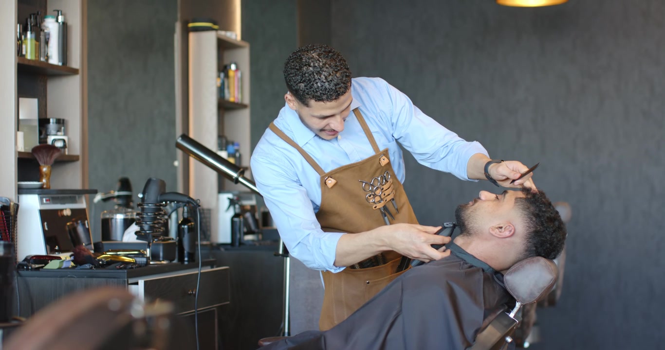 Mandlig barber i forklæde trimmer og former skæg på en mandlig kunde, der læner sig tilbage, ved hjælp af en trimmer i barbersalonen