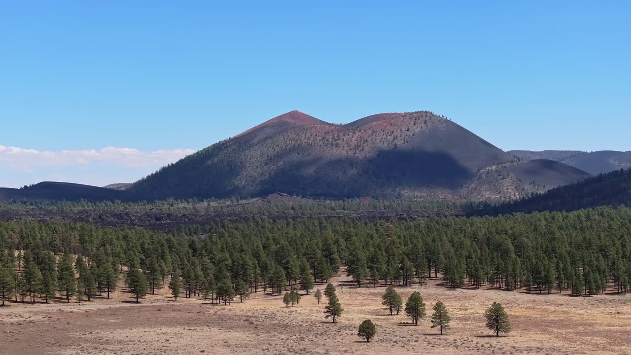 Un primer plano aéreo con dron que se eleva y revela el Monumento Nacional del Volcán Sunset Crater cerca de Flagstaff, Arizona, mientras los árboles y un campo de hierba pasan frente al cono de ceniza en un día soleado