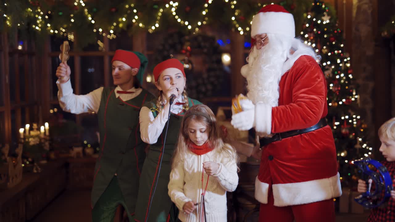 Christmas celebration with Santa. Fairy elves, Santa and children are singing and dancing on Christmas background indoors.