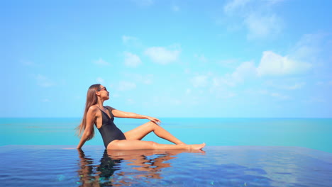 Mujer-Exótica-Sexy-En-Traje-De-Baño-Sentada-En-El-Borde-De-La-Piscina-Infinita-Disfrutando-De-Una-Vista-Impresionante-Del-Horizonte-Del-Mar-Tropical,-Cámara-Lenta