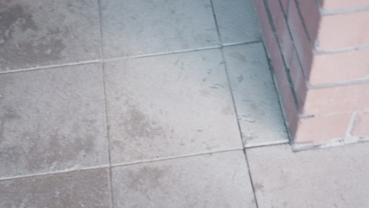 el marco inferior muestra a una mujer con un abrigo oscuro subiendo una escalera junto a un poste de ladrillo y una barandilla de hierro, un bolso marrón balanceándose, enfoque en la vista del pasillo, movimiento capturado desde arriba con énfasis en las piernas y el movimiento