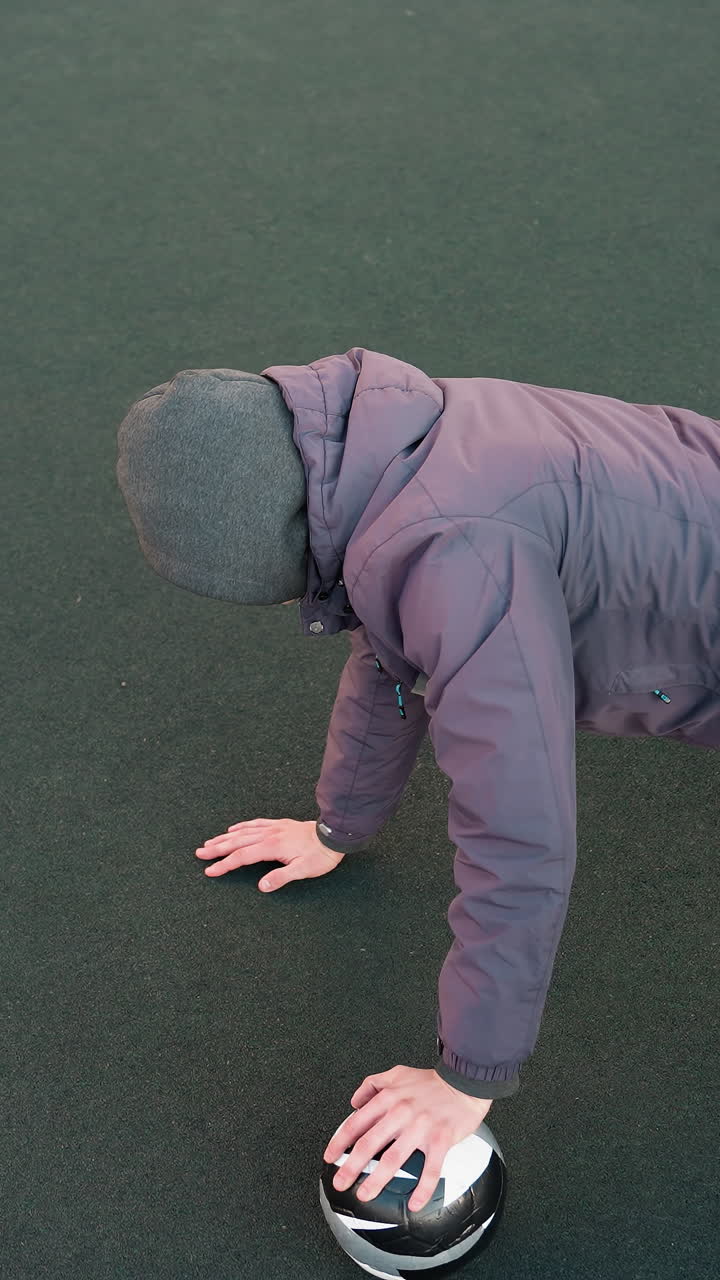 평평한 녹색 표면에서 야외 훈련 기간 동안 힘, 통제 및 민성을 보여주는 축구 공에 손을 교차하여 푸시 업을 수행하는 운동 선수의 오버 헤드 뷰