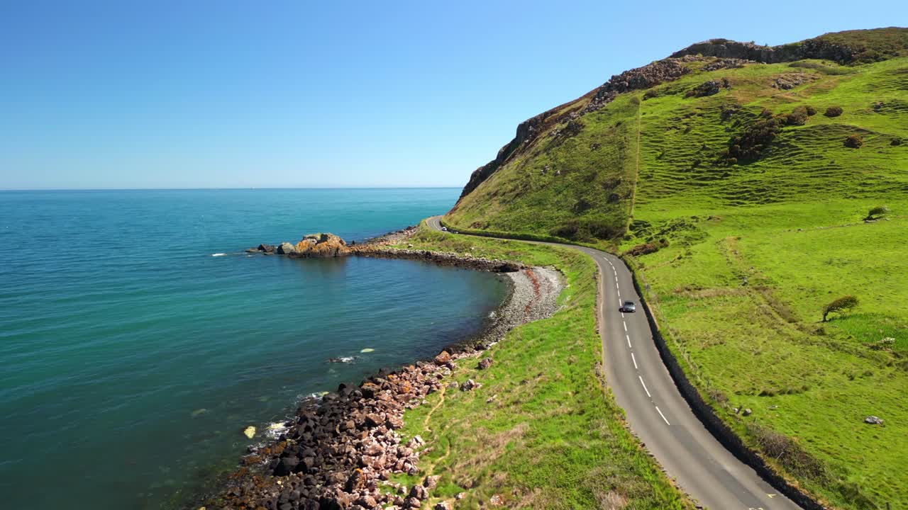 Aerial video of the Antrim Coastal Route and travelling cars on a bright and sunny day. The blue sea and the green cliffs pass by the camera. Filmed in Ballygally in 4K, 60FPS and with Rec709 color.