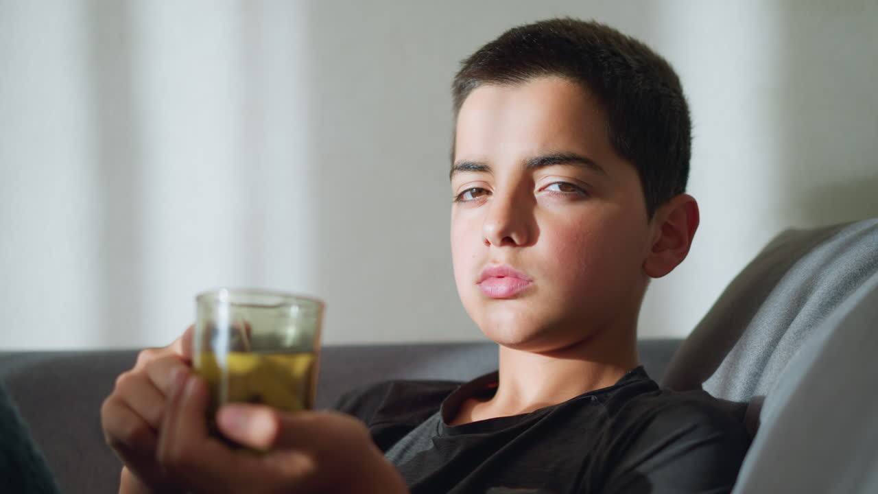 Boy with tea cup staring at camera, holding cup close to his mouth while looking tired and sick, with sunlight reflecting off his face, creating a soft, warm ambiance