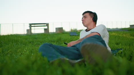 young man enjoying music outdoors, lying on his side on a grassy field, wearing headphones, looking down at his smartphone, with a blurred background featuring a bench and distant scenery