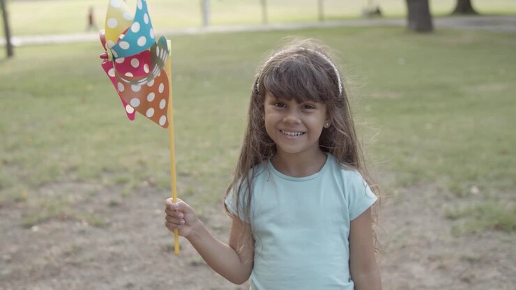 Menina latina segurando um leque de papel, parada no parque e sorrindo para a câmera