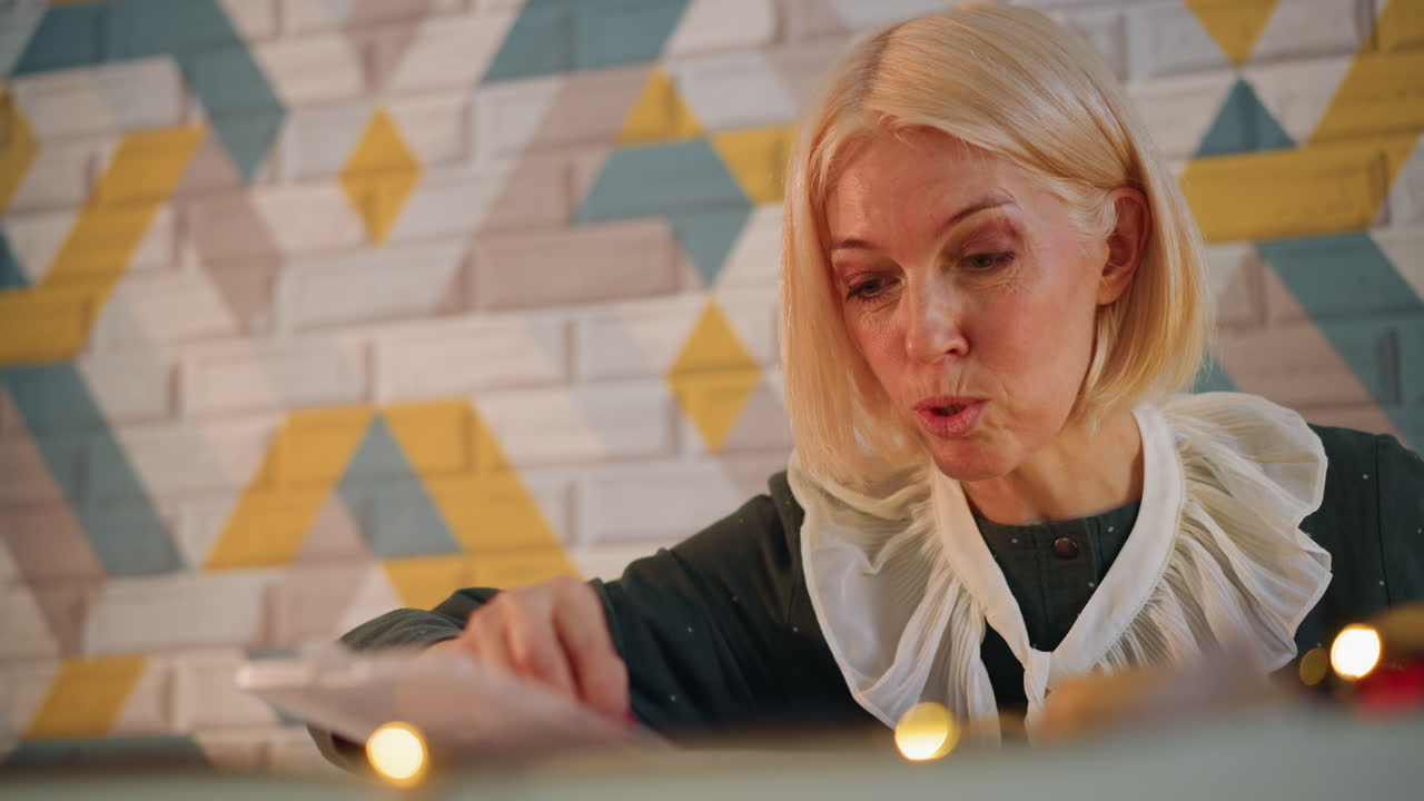 Blonde Frau schreibt Weihnachtskarte bei Lichterketten, versiegelt Umschlag, lächelt sanft, gemusterter Fliesen-Hintergrund, warmes Bokeh, Geschenkanhänger und Schreibtisch-Schreibwaren, intime Feiertagsatmosphäre, sanfter Fokus