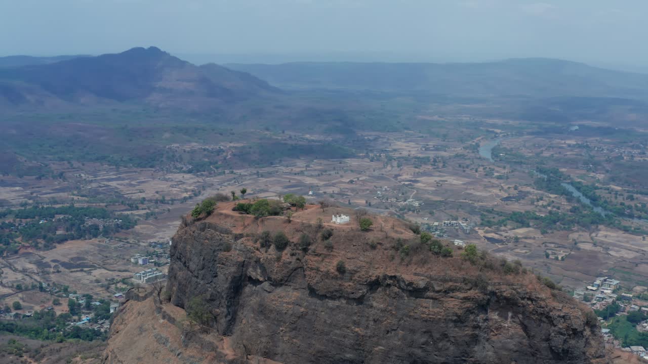 High aerial of Sarasgad Fort's plateau top with small temple amid vast dry valley landscape, distant river, hills, sparse trees, and expansive Raigad plains