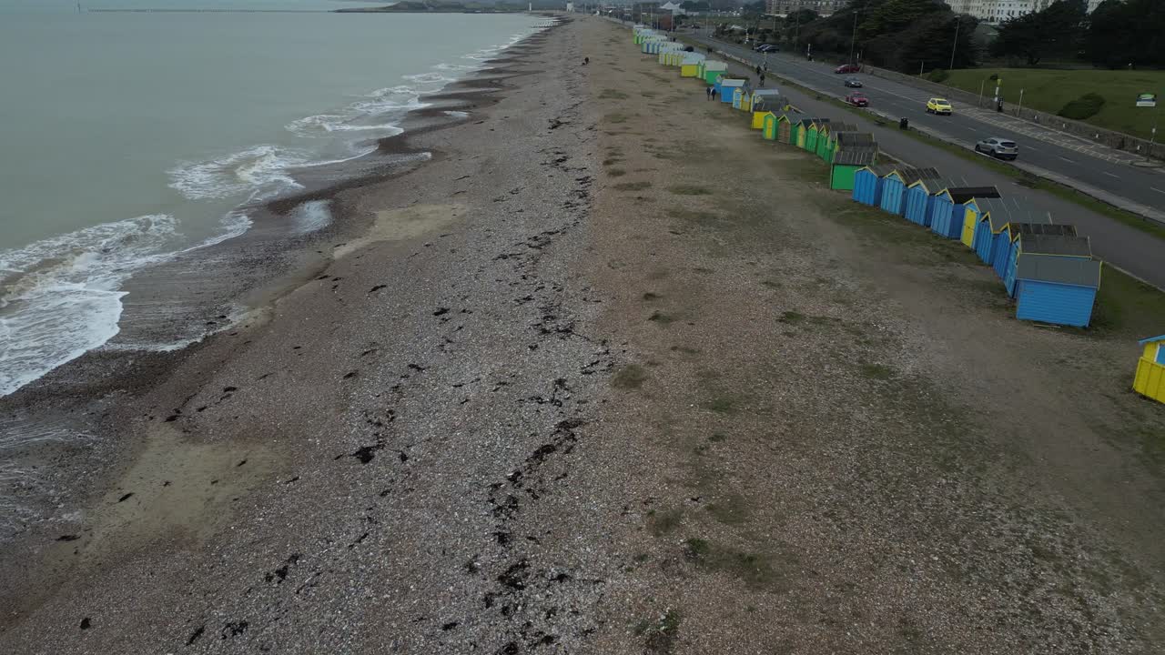 Overcast day on the UK coast. Gentle waves meet the shingle beach next to a road with moving cars. Colorful beach huts line the shore. A typical British seaside view.