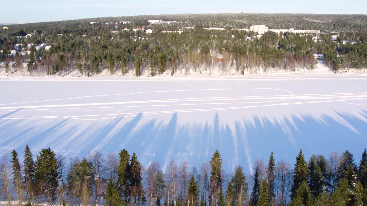 Una strada panoramica fiancheggiata da laghi ghiacciati sotto un tramonto a Rovaniemi, Lapponia finlandese. Le ombre dei pini creano motivi suggestivi mentre il drone avanza attraverso il paesaggio artico.