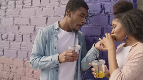 young couple sharing a tasty hamburger and drinking a cold drink together, while chatting leaning against a colourful brick wall