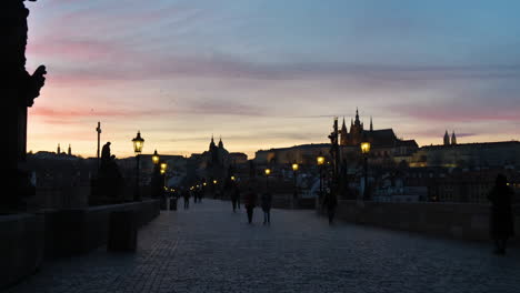 Oscuridad-Y-Siluetas-De-Personas-En-El-Puente-Charles,-Praga-República-Checa-Con-Castillo-En-La-Colina-Bajo-El-Cielo-Del-Atardecer-Durante-La-Pandemia-Del-Virus-Covid-19,-Cámara-Lenta