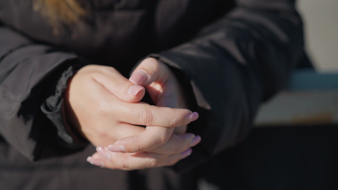 Mani di donna con unghie lunghe e curate, giunte in un gesto rilassato. L'illuminazione soffusa evidenzia la texture della pelle e i dettagli delle unghie su uno sfondo di abiti scuri. Ambiente esterno