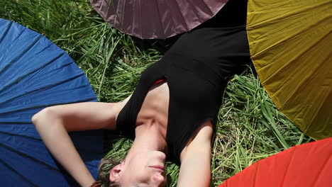A woman lays out on the grass in the sun surrounded by for colored umbrellas