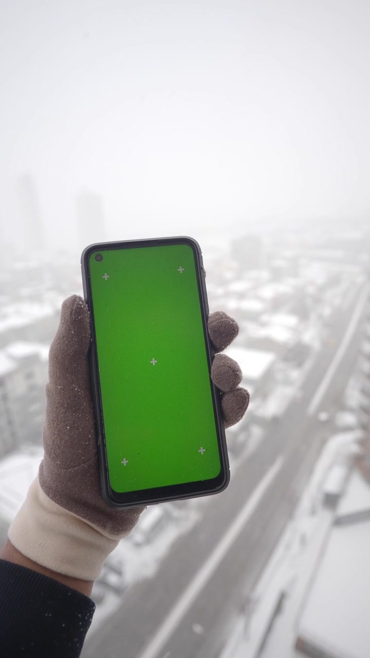 Behandschuhte Hand hält Smartphone mit Greenscreen in verschneiter Stadt