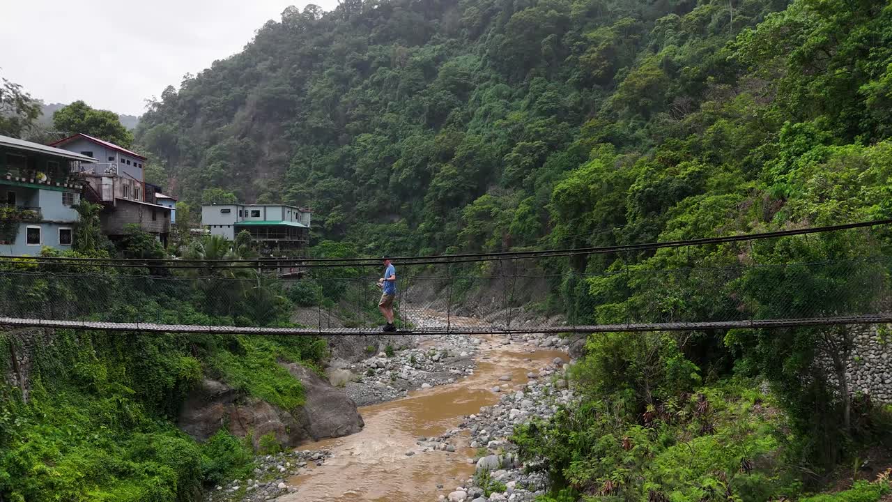 The footage showcases a stunning view of a valley in the mountains, and a man strolling across a suspension bridge near bridal falls outside of baguio