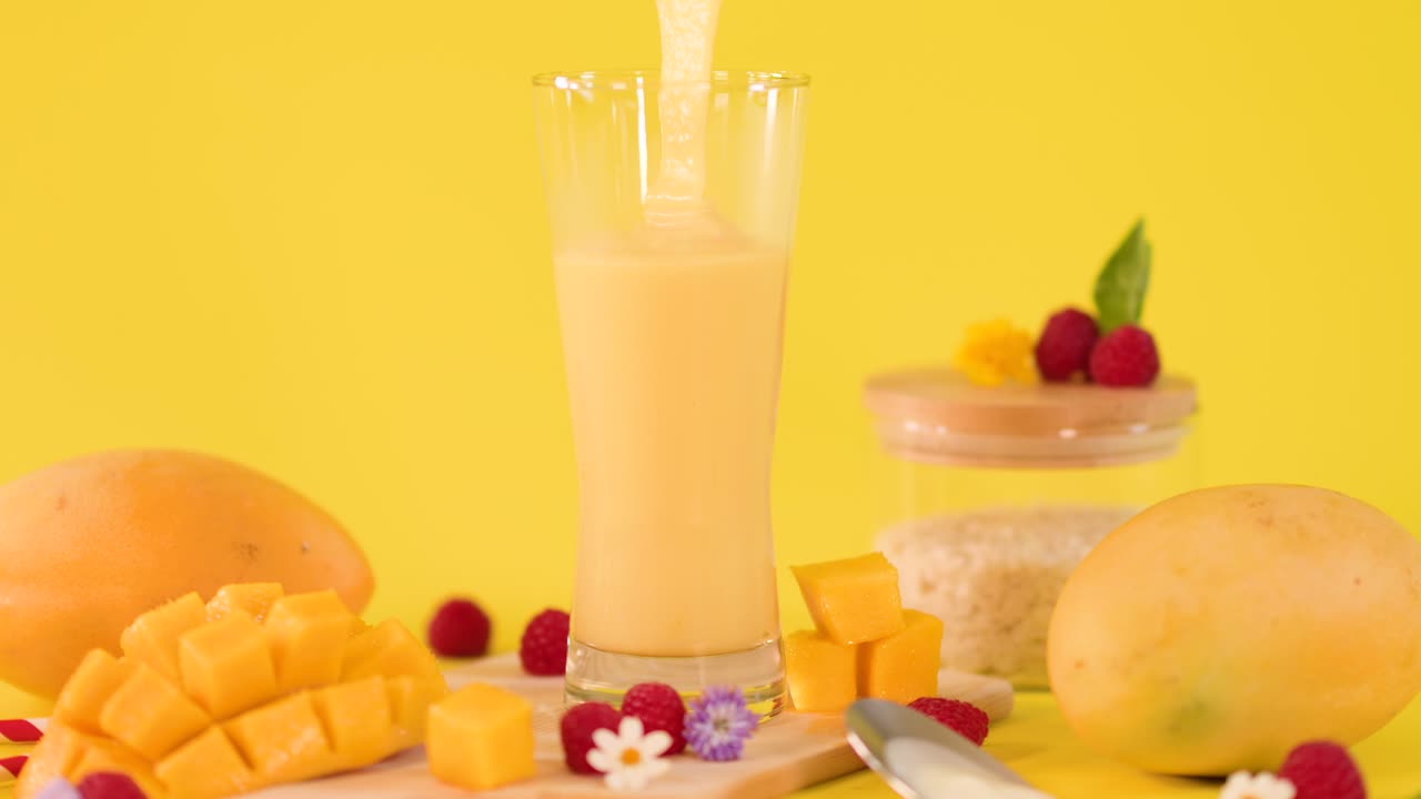 Smoothie cremoso al mango versato in un bicchiere alto tra frutta fresca su sfondo giallo brillante