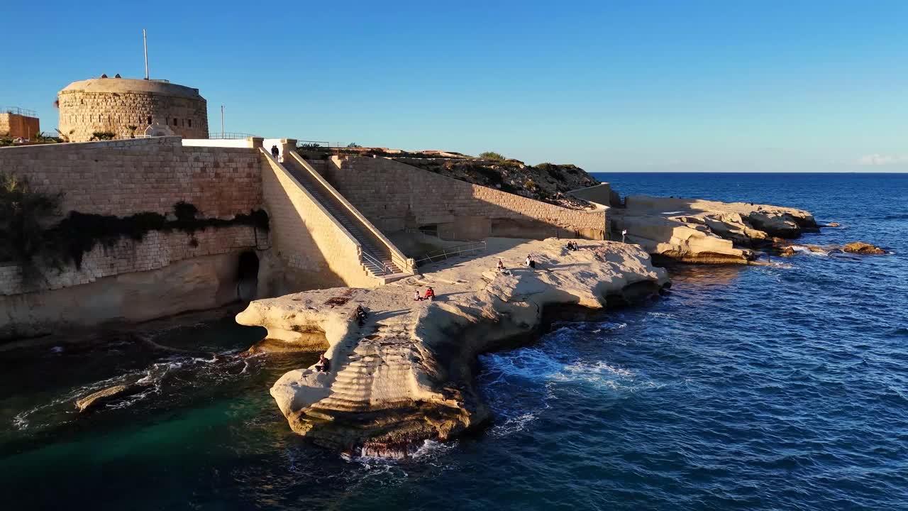 Una ripresa aerea con drone rivela le storiche mura in pietra di Forte Tigne a La Valletta, Malta, affacciate su una costa rocciosa con scale scolpite che conducono al Mar Mediterraneo.